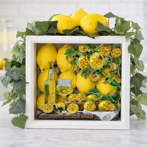 May include: A white framed display featuring bright yellow lemons, a bottle, and a glass, all adorned with yellow embellishments. Green ivy frames the top and sides, creating a vibrant, summery composition. The display is set on a white surface.
