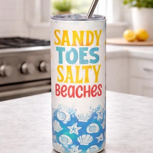 May include: A white tumbler with a silver straw, featuring the text "SANDY TOES SALTY BEACHES" in yellow, blue, and red. The bottom half has a blue wave design with seashell and starfish illustrations. A beach-themed drinkware item.