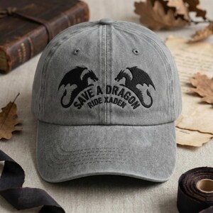 May include: A gray baseball cap with a curved brim. The cap features a black dragon graphic and the text "SAVE A DRAGON RIDE XADEN" in black. The hat is made of a soft, textured fabric.