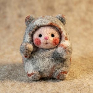 May include: A handmade felted doll in a gray bear hoodie. The doll has a pink face with rosy cheeks and small black eyes. The bear hoodie has small ears and red accents. The doll is standing on a beige surface.
