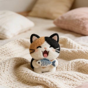 May include: A handcrafted felt cat figurine in calico colors, holding a blue fish. The cat has a happy expression with its mouth open. The figurine is set on a cream-colored knitted blanket.