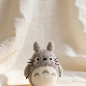Puede incluir: Una figura gris de Totoro con vientre blanco y mejillas rosas. La figura tiene orejas peque&ntilde;as, bigotes negros y ojos grandes y redondos. Est&aacute; sobre una superficie blanca y texturizada.