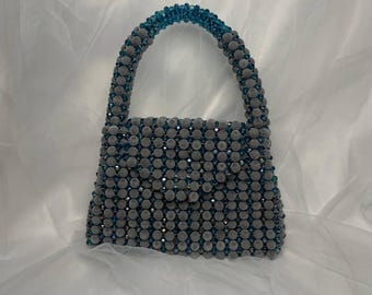 May include: A gray beaded handbag with a rounded handle and a rectangular body. The handle and top edges feature teal beads. The bag has a button closure and is set against a white background.