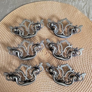 Könnte beinhalten: Sechs antike, silberfarbene Schubladengriffe mit aufwendigen, scrollartigen Designs. Jeder Griff verfügt über einen gebogenen Griff und dekorative Verzierungen, ideal, um Möbeln einen Vintage-Touch zu verleihen. Die Griffe sind auf einer gewebten, hellbraunen Oberfläche angeordnet.