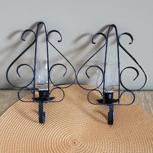 May include: A pair of black metal wall sconces featuring a decorative scrollwork frame and a clear glass candle holder cup. Each sconce has a pointed top and a curved base, designed to hold a single candle within the central glass vessel.