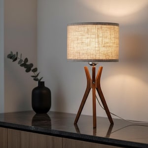 May include: A wooden tripod table lamp with a beige linen shade. The lamp has a warm, inviting glow and sits on a dark countertop. A black vase with greenery is to the left of the lamp.