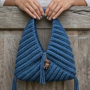 Può includere: Una borsa blu all'uncinetto fatta a mano con una forma triangolare unica. La borsa presenta un motivo a punto strutturato e un piccolo ciondolo con una nappa. La borsa è tenuta da una persona.