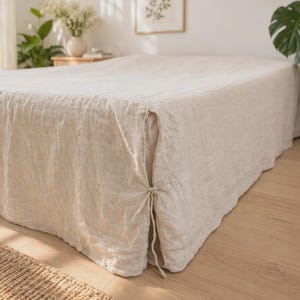 May include: A beige linen bed cover draped over a mattress, featuring a textured fabric surface and side panels that are secured at the corners with thin, matching fabric ties that hang down along the side of the bed frame.