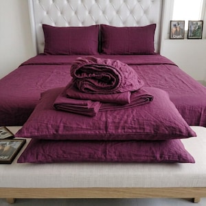 May include: A deep plum-coloured bedding set featuring a flat sheet, a fitted sheet, and two pillowcases. The set is displayed on a bed with a tufted white headboard, with the linens arranged in a folded stack on top of the bed surface.
