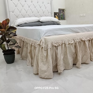 Può includere: Un letto con testiera bianca trapuntata, cuscini bianchi e grigi e un lenzuolo bianco. La base è coperta da una balza da letto beige con bordo arricciato. Una pianta in vaso con foglie variegate verdi e rosse si trova a sinistra del letto.