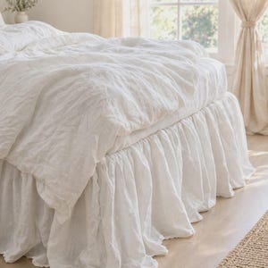 May include: A white bed skirt with a gathered, ruffled design that extends to the floor. The fabric has a textured, slightly wrinkled appearance and is draped around the base of a bed frame, providing a soft, layered look to the bedding.