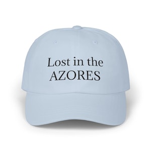 May include: A light blue baseball cap featuring the black text Lost in the AZORES embroidered across the front panel. The cap has a structured crown, a curved brim, and a button on the top center, providing a simple and clean design aesthetic.