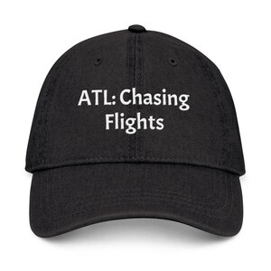 Puede incluir: Gorra de béisbol negra con el texto blanco "ATL: Chasing Flights". La gorra tiene una visera curva y un botón en la parte superior. Un accesorio de viaje.