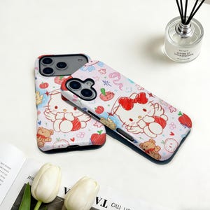 Puede incluir: Dos fundas de tel&eacute;fono blancas y rosas con un personaje de gato de dibujos animados, un lazo rojo, alas de &aacute;ngel y un osito de peluche. Las fundas tienen un patr&oacute;n repetido de manzanas, flores y corazones. Las fundas est&aacute;n sobre una superficie blanca.