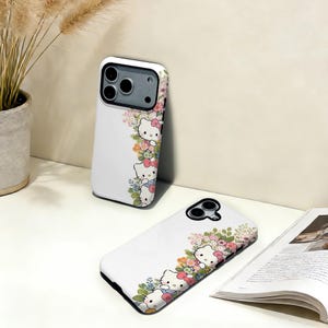 Puede incluir: Fundas blancas para tel&eacute;fono con un dise&ntilde;o floral y de gato de dibujos animados. Las fundas presentan un patr&oacute;n repetido del personaje del gato con lazos rosas y flores coloridas. Las fundas se muestran sobre una superficie blanca.