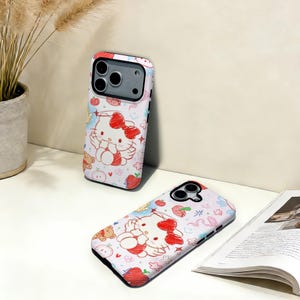 Puede incluir: Dos fundas blancas para tel&eacute;fono con un dise&ntilde;o colorido que presenta un gato de dibujos animados con un lazo rojo, alas de &aacute;ngel y otras ilustraciones lindas. Las fundas tienen un borde rojo y est&aacute;n colocadas sobre una superficie blanca.