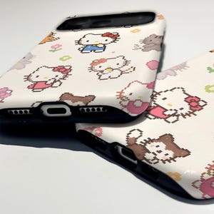 May include: Two white phone cases with a pixelated design featuring Hello Kitty and other characters. The cases have a black border and are decorated with pink, brown, and blue accents. The cases are likely for a smartphone.