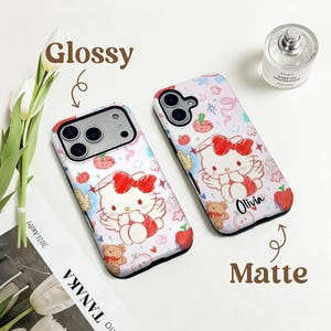 Puede incluir: Dos fundas de tel&eacute;fono con fondo blanco con un gato de dibujos animados con un lazo rojo, alas de &aacute;ngel y un osito de peluche. Una funda es brillante y la otra mate. La palabra "Olivia" est&aacute; escrita en una funda.