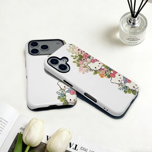 Puede incluir: Fundas blancas para tel&eacute;fono con dise&ntilde;o floral y personajes de Hello Kitty. Las fundas tienen un acabado brillante y un recorte negro para la c&aacute;mara. El dise&ntilde;o incluye elementos florales rosas, blancos y verdes.