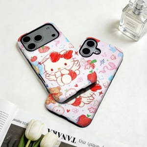 Puede incluir: Dos fundas para tel&eacute;fono con fondo blanco. Las fundas presentan un dise&ntilde;o de dibujos animados de un gato blanco con un lazo rojo, alas y otras ilustraciones. Las fundas tienen un borde negro y est&aacute;n decoradas con manzanas rojas, ositos de peluche y otros elementos lindos.