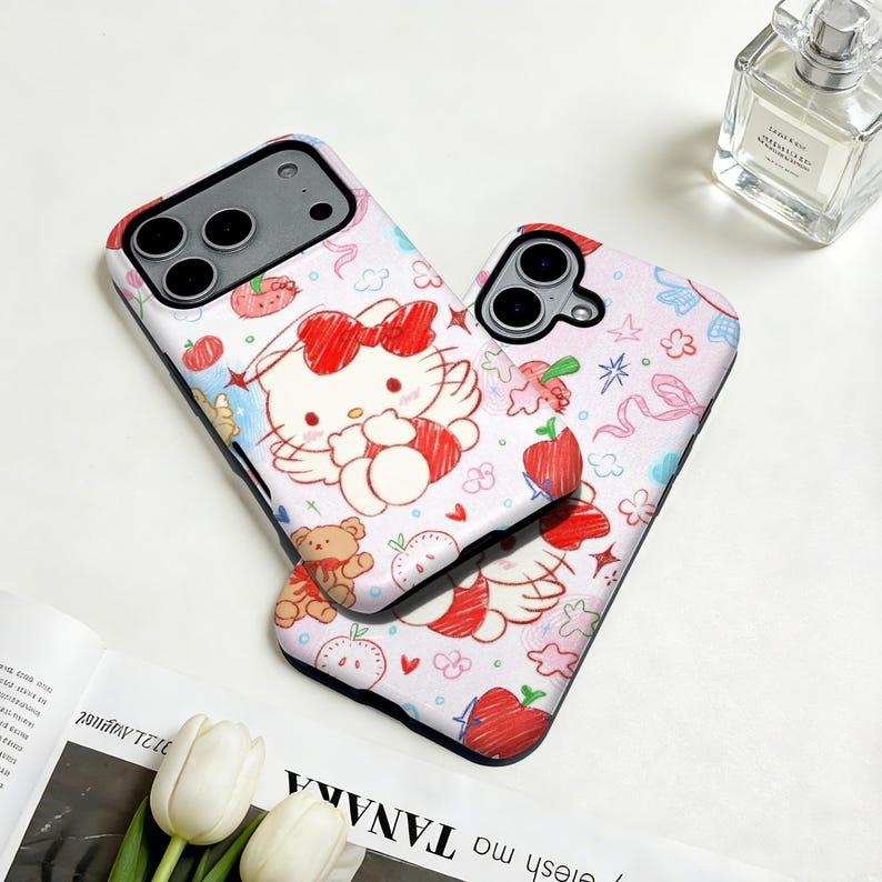 Puede incluir: Dos fundas para tel&eacute;fono con fondo blanco. Las fundas presentan un dise&ntilde;o de dibujos animados de un gato blanco con un lazo rojo, alas y otras ilustraciones. Las fundas tienen un borde negro y est&aacute;n decoradas con manzanas rojas, ositos de peluche y otros elementos lindos.