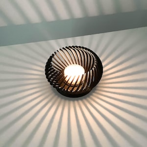 May include: A black, modern wall sconce with a spiral design. The light fixture has a central bulb, casting a warm glow and creating a pattern of shadows on the wall. The sconce is mounted on a white wall.