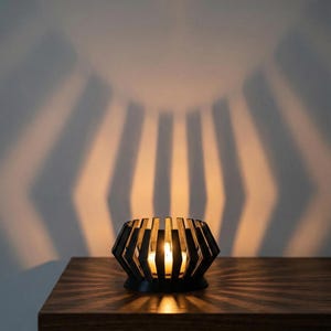 May include: A black geometric candle holder with a lit candle inside, casting striped shadows on the wall. The candle holder sits on a wooden surface, creating a warm, inviting ambiance. The design is modern and minimalist.