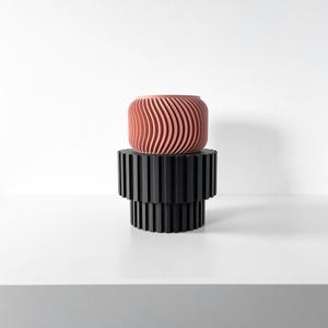 Può includere: Un moderno supporto nero a due livelli sostiene un vaso cilindrico color terracotta. Il vaso presenta un motivo a onde. Il supporto ha un design a forma di ingranaggio. Gli oggetti sono posti su uno sfondo bianco.