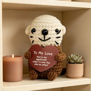 Könnte beinhalten: Ein gehäkeltes Otter-Plüschtier mit cremefarbenem Kopf und braunem Körper hält ein herzförmiges Schild mit der Aufschrift "To My Love". Eine brennende Kerze und eine kleine Topf-Sukkulente befinden sich ebenfalls auf dem Regal.