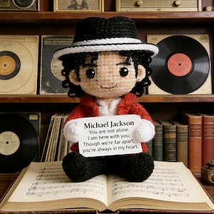 Puede incluir: Muñeco de ganchillo de Michael Jackson con sombrero negro y chaqueta roja, sosteniendo un cartel con un mensaje. El muñeco está sentado sobre un libro de partituras abierto, con discos de vinilo y libros al fondo.