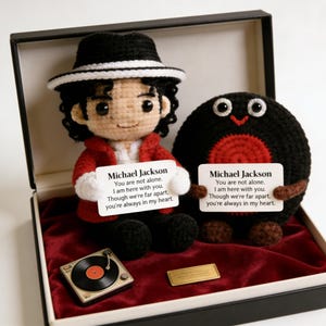 Könnte beinhalten: Gehäkelte Michael Jackson-Puppe und eine runde, schwarze und rote Plüschfigur, beide halten Karten mit Text. Präsentiert in einer schwarzen Box mit rotem Samtboden, zusammen mit einem Miniatur-Plattenspieler und einer goldenen Plakette.