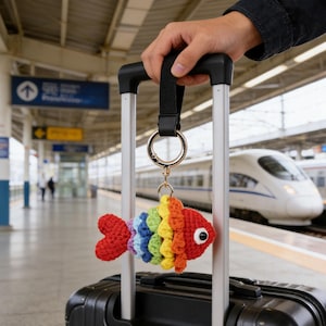Op de afbeelding: Een kleurrijke, gehaakte vissen sleutelhanger met een regenboogontwerp, bevestigd aan een zwarte koffer. De vis heeft rode, oranje, gele, groene, blauwe en paarse schubben. De sleutelhanger wordt vastgehouden door een zwarte band en een gouden ring.