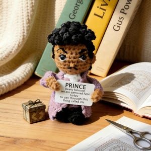 May include: A handmade crochet doll of Prince, holding a sign with lyrics. The doll has a purple coat and black hair. A small decorative box and books are in the background. The sign reads "PRINCE Dearly beloved, we are gathered here today to get through this thing called life."