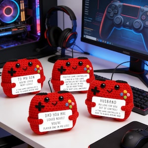 Op de afbeelding: Vier rode gehaakte gamecontrollers met zwarte kruis- en kleurrijke knopdetails. Elke controller houdt een wit bord vast met een bericht: "To My Son", "Positive Game Controller!", "Dad You Are Loved" en "Husband". Een gaming-opstelling staat op de achtergrond.