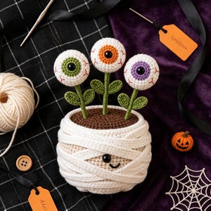 May include: A crocheted mummy-themed planter pot featuring three eyeball flowers with green, orange, and purple irises. The pot is wrapped in white yarn strips, has a small black eye and a stitched smile, and contains brown soil-coloured yarn.