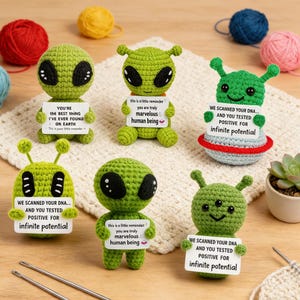 May include: A collection of handmade green crochet alien figures, each holding a small sign with positive messages. The aliens have large black eyes and antennae. Yarn and crochet hooks are in the background.