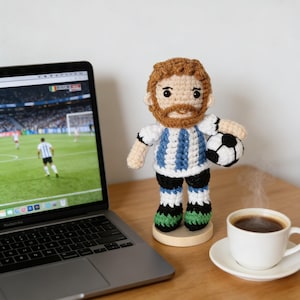 May include: A handmade crochet doll of a soccer player wearing a blue and white striped jersey, black shorts, and green and black socks, holding a soccer ball. The doll is on a wooden base next to a laptop and a cup of coffee.