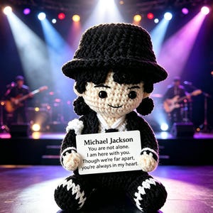 Könnte beinhalten: Gehäkelte Puppe von Michael Jackson mit schwarzem Hut, Jacke und Hose, die ein Schild mit einer Botschaft hält. Die Puppe befindet sich auf einer Bühne mit Musikern im Hintergrund, beleuchtet von Bühnenlichtern.