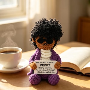 May include: A crocheted Prince doll with sunglasses, a purple jacket, and a white shirt, holding a sign saying "EMOTIONAL SUPPORT PRINCE." A cup of coffee and an open book are on the table.