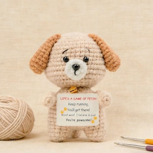 May include: A crocheted beige dog with brown ears and a small gold bell. The dog holds a sign that reads "LIFE'S A GAME OF FETCH" with encouraging phrases. A ball of yarn and crochet hooks are in the background.