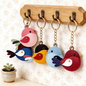 May include: A collection of five bird-shaped keychains in various colors, including pink, blue, yellow, and red. Each keychain has a gold-colored chain and a metal ring for attaching keys. The keychains are hanging on a wooden rack.
