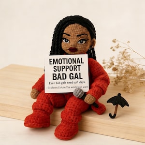May include: A handmade crochet doll in a red outfit, holding a sign that reads "EMOTIONAL SUPPORT BAD GAL." The doll has black hair, a microphone, and a small black umbrella. The text on the sign says, "Even bad gals need soft days. -Sit down. Exhale. The world can wait."
