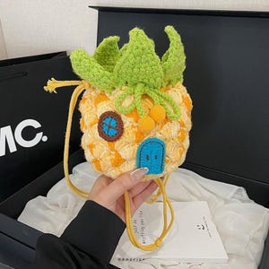 May include: A yellow and orange crocheted pineapple-shaped bag with green leaves and a yellow drawstring. The bag features a blue door and a window with a blue cross design. The bag is held by a person.