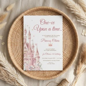 May include: A princess-themed invitation card with the text "One-ce Upon a time..." and "Princess Olivia". The card features a pink castle illustration and floral accents. The invitation is set on a woven tray with decorative dried plants.