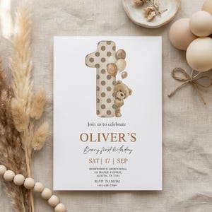 Pode incluir: Um cart&atilde;o de convite branco com o texto "Join us to celebrate OLIVER'S Beary first birthday". O n&uacute;mero um &eacute; decorado com bolinhas marrons e um ursinho de pel&uacute;cia segurando bal&otilde;es. O texto adicional inclui a data, o local e as informa&ccedil;&otilde;es RSVP.
