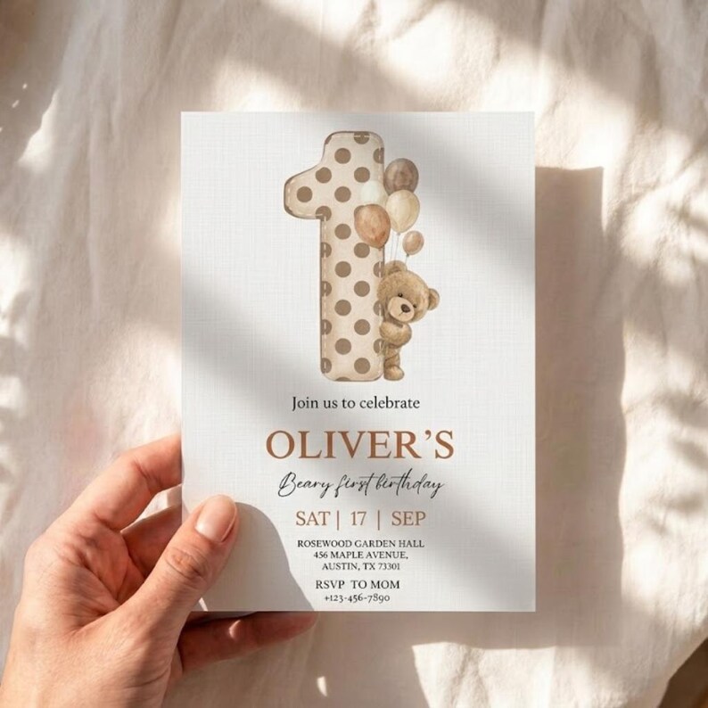 Pode incluir: Um convite de anivers&aacute;rio bege com o texto "Join us to celebrate OLIVER'S Beary first birthday". O design inclui um n&uacute;mero um com bolinhas, um ursinho de pel&uacute;cia e bal&otilde;es. O convite &eacute; segurado por uma m&atilde;o.