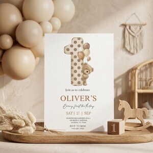Pode incluir: Um convite de anivers&aacute;rio com um ursinho segurando bal&otilde;es e o n&uacute;mero um. O convite diz "Beary first birthday de Oliver" com a data e o local. Um cavalo de balan&ccedil;o de madeira e um bloco com o n&uacute;mero um tamb&eacute;m s&atilde;o vis&iacute;veis.