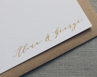 May include: A white card with gold lettering that reads "Alina & George". The card is folded in half and the top half is resting on a brown envelope.