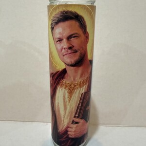 May include: A tall, cylindrical glass prayer candle featuring a printed image of a man with short brown hair and light stubble. He wears a white tunic with gold trim and a red cloak, holding a dark book. A golden halo glows behind his head.