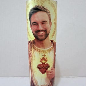 May include: A clear glass prayer candle featuring a custom photographic print of a smiling person with a beard, depicted in the style of the Sacred Heart of Jesus with a golden halo, a white tunic, and a flaming heart encircled by thorns.